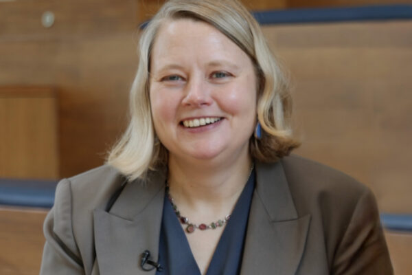 Elinor Karlsson, chief scientist of Darwin's Ark, wears a brown blazer and blue top and sits in a wood-paneled room at the Broad Institute of MIT and Harvard. She is smiling at the camera, conveying a sense of both intelligence and approachability.